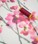 Seville Blossom Stone floral fabric photographed with a burgundy thread spool for scale, highlighting its painterly pink blossoms, raspberry accents and grey-green branches on a neutral stone background.