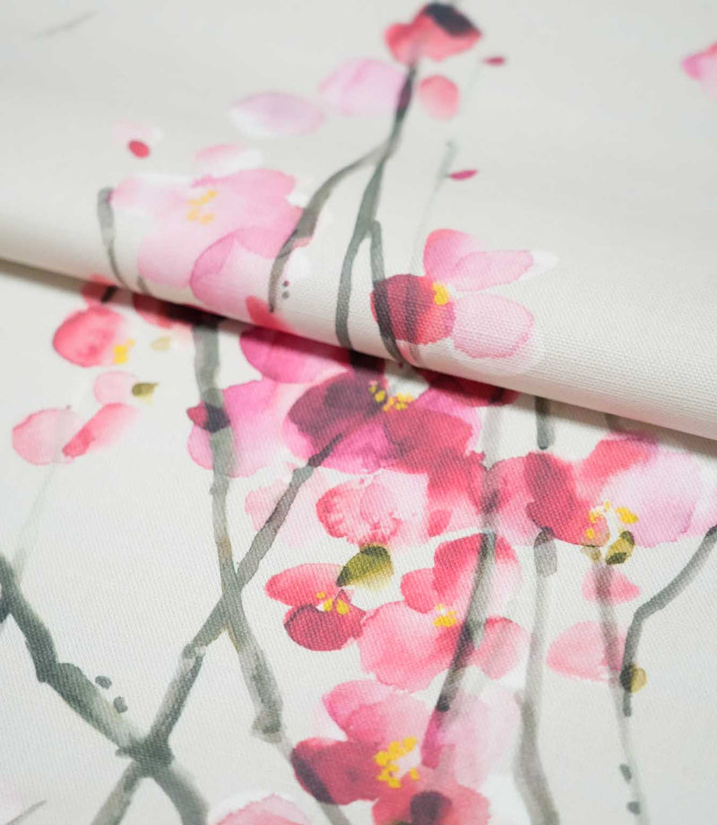 Seville Blossom Stone fabric arranged in a soft fold, displaying clusters of pink and deep raspberry watercolour blossoms with yellow centres and slender grey-green branches.
