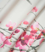 Seville Blossom Stone fabric gathered into flowing folds, showing its natural drape and delicate floral pattern of pink and raspberry blossoms scattered across grey-green branches on a soft stone background.