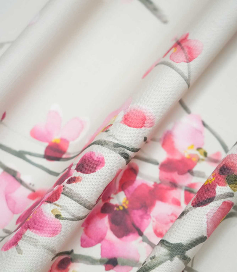 Seville Blossom Stone fabric gathered into flowing folds, showing its natural drape and delicate floral pattern of pink and raspberry blossoms scattered across grey-green branches on a soft stone background.