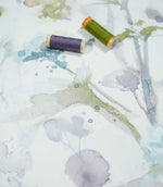 Sorong Lemon floral fabric photographed with purple and olive green thread spools for scale, highlighting its delicate blue, lavender and yellow-green watercolour flowers on a soft off-white background.