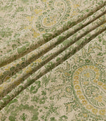 Southold Spring fabric draped in soft diagonal folds, showcasing its detailed green and yellow paisley floral pattern, delicate trailing foliage and textured cream background, with the folds highlighting the fabric's natural drape and intricate design.