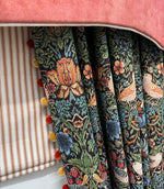 Close-up of Strawberry Thief Black fabric curtains featuring intricate birds, strawberries and botanical foliage in green, blue, orange and red, finished with decorative pom-pom trim beneath a coral pelmet.
