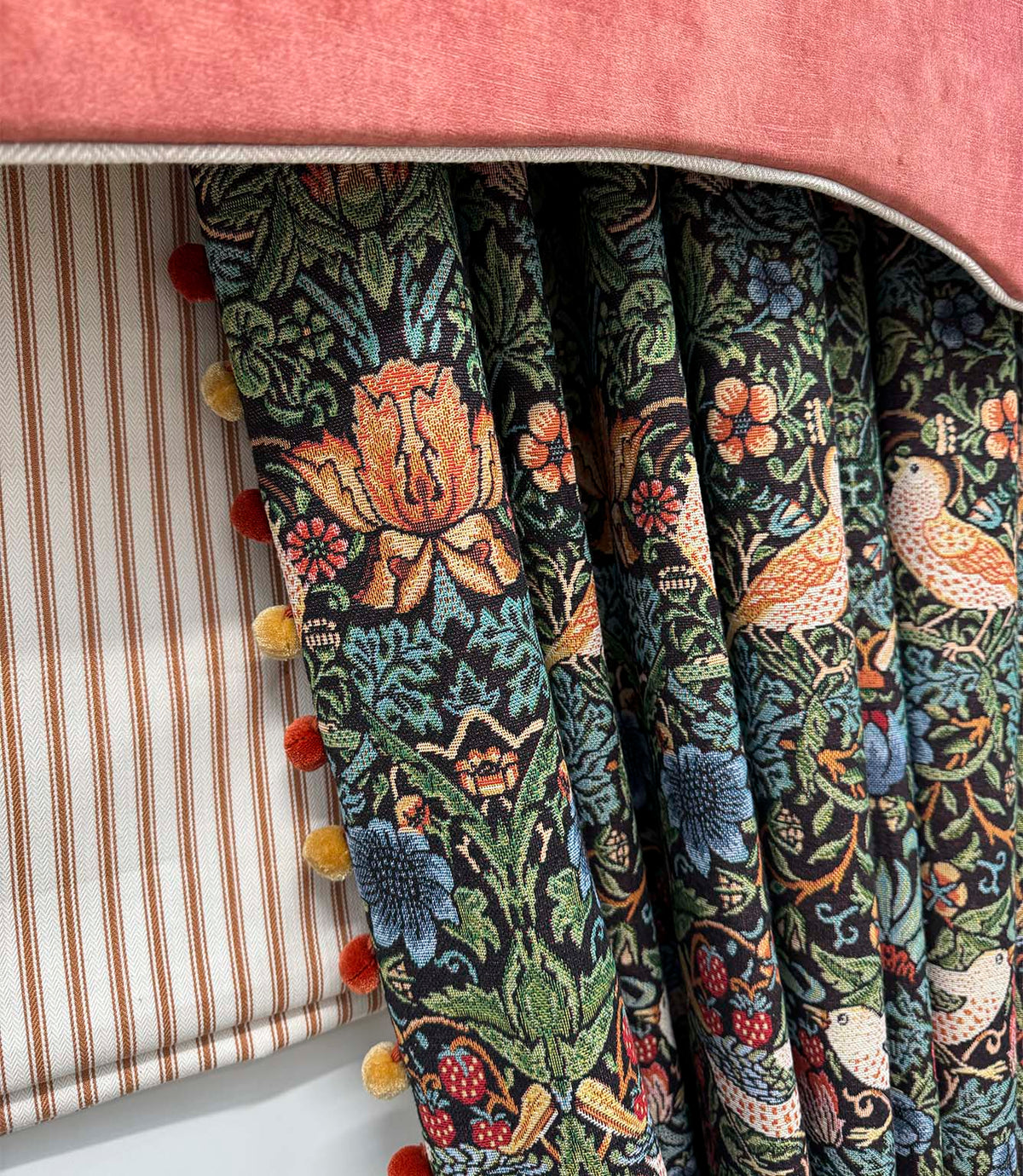 Close-up of Strawberry Thief Black fabric curtains featuring intricate birds, strawberries and botanical foliage in green, blue, orange and red, finished with decorative pom-pom trim beneath a coral pelmet.