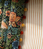 Close-up of Strawberry Thief Tapestry Noir curtain fabric with an intricate bird and floral design, finished with red and mustard pom-pom trim beside a cream and rust striped Roman blind.