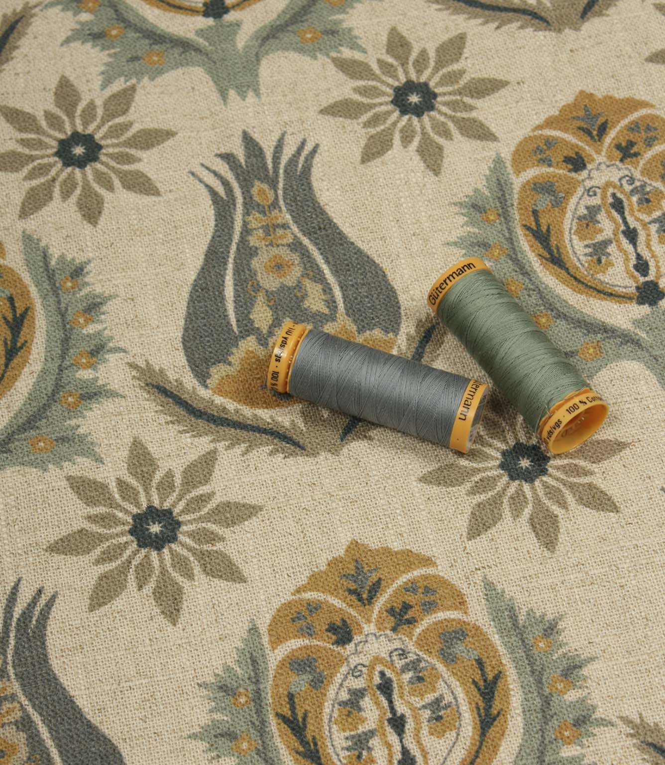 Decorative botanical fabric shown with coordinating sage green and blue thread reels on a natural linen-look ground.