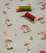 Toadstool Meadow Multi woodland fabric photographed with green and burgundy sewing thread spools for scale, showing the small red and brown mushroom illustrations and scattered fern motifs on a natural woven surface.