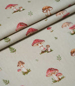 Toadstool Meadow Multi fabric arranged in overlapping folds, featuring a repeating woodland pattern of red and brown toadstools, small green fern sprigs and detailed mushroom illustrations on a beige woven surface.