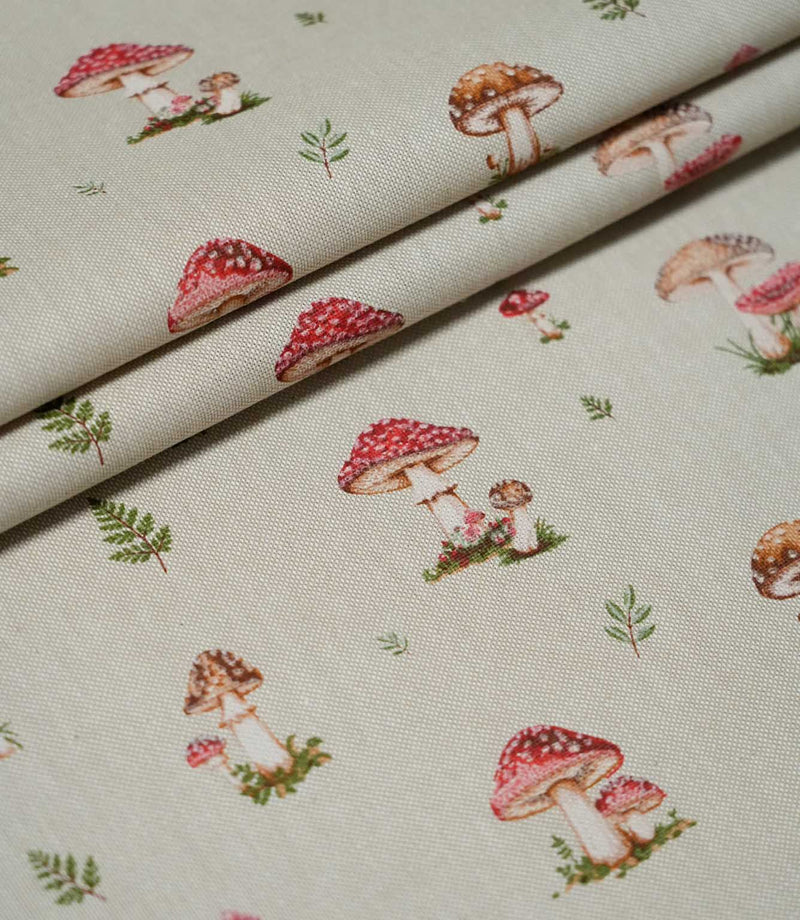 Toadstool Meadow Multi fabric arranged in overlapping folds, featuring a repeating woodland pattern of red and brown toadstools, small green fern sprigs and detailed mushroom illustrations on a beige woven surface.