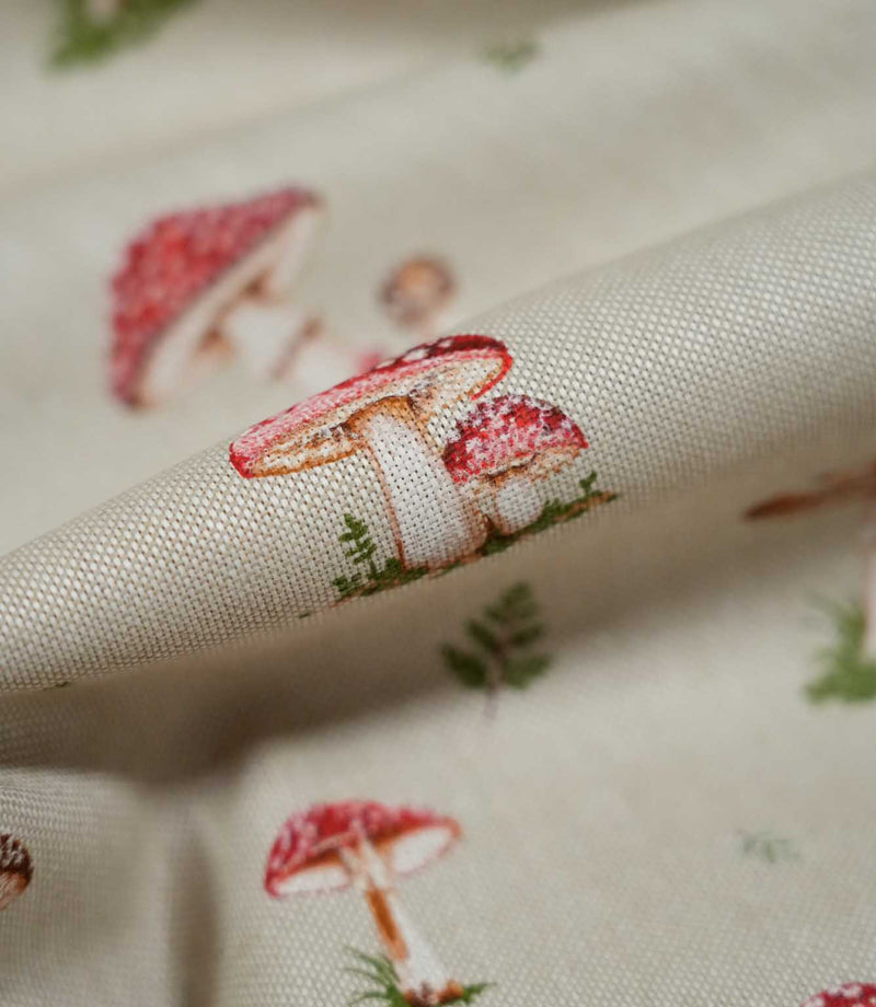 Detailed close-up of Toadstool Meadow Multi fabric showing two red and pink spotted woodland mushrooms with cream stems and green foliage, printed onto a textured natural beige woven surface.