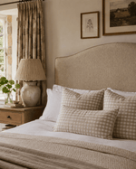 A warm Cotswold-inspired bedroom with a curved headboard upholstered in textured beige woven fabric, styled with neutral gingham pillows, layered bedding, soft natural light and relaxed rustic furnishings.