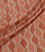 Wading Coralee fabric arranged in soft diagonal folds, showcasing its contemporary wave-inspired pattern in coral, terracotta and muted pink, with delicate cream scalloped edges, fine brown detailing and a subtly textured woven appearance.