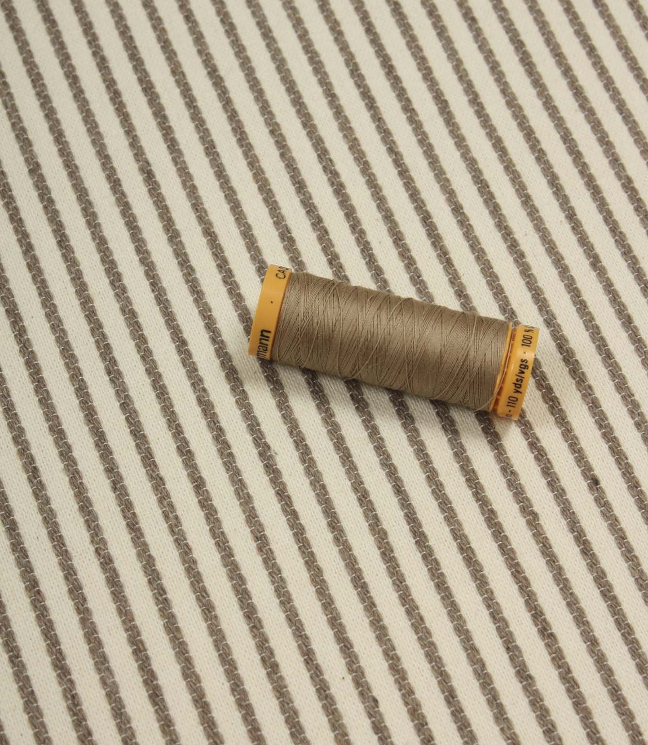 Wedmore Wide ivory striped fabric displayed with coordinating taupe thread spool, highlighting the fine embroidered stripe detail.
