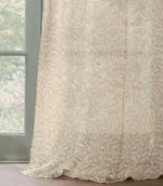 Full-length Pure Willow Boughs sheer curtain in a neutral linen beige and ivory botanical design, softly draping onto a wooden floor beside a green framed window.