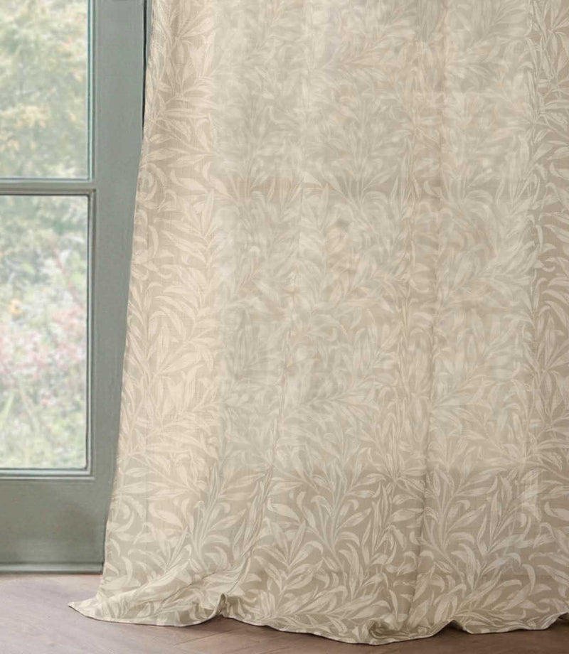 Full-length Pure Willow Boughs sheer curtain in a neutral linen beige and ivory botanical design, softly draping onto a wooden floor beside a green framed window.