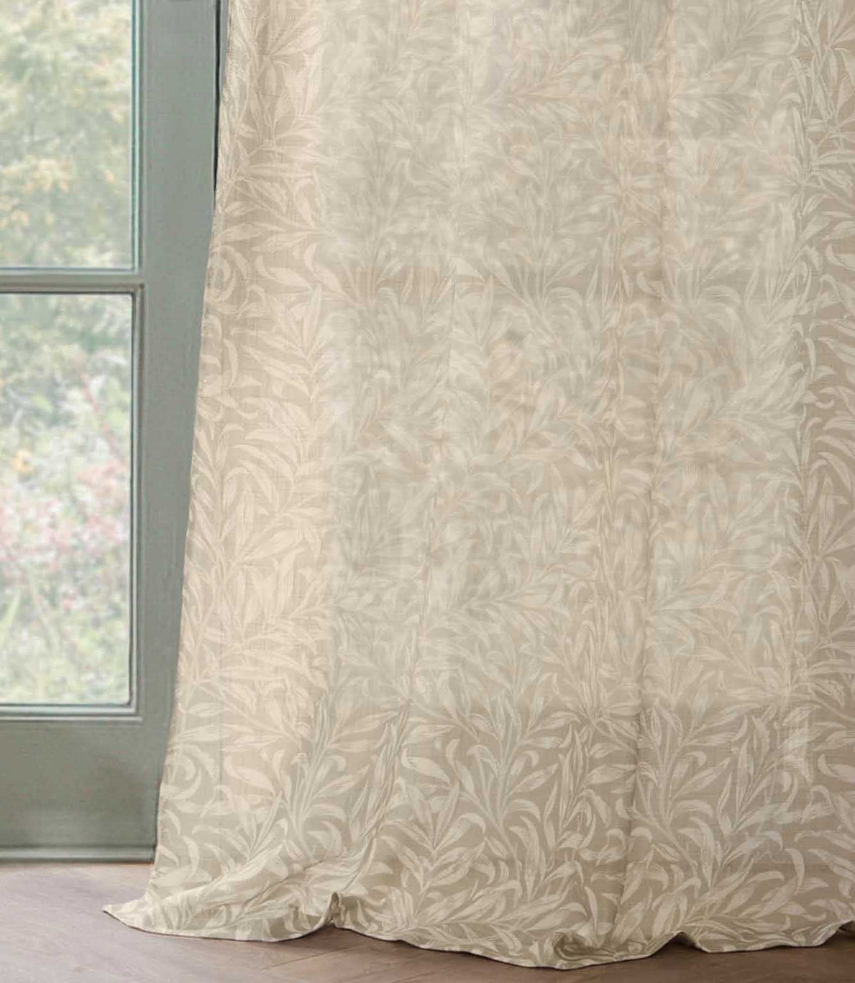 Full-length Pure Willow Boughs sheer curtain in a neutral linen beige and ivory botanical design, softly draping onto a wooden floor beside a green framed window.