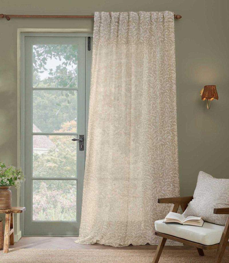 William Morris Pure Willow Boughs sheer curtain in linen beige and ivory, hanging at a tall green framed window in a calm living room with natural wood furniture.