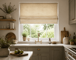 A warm, premium country-style kitchen interior with soft natural daylight streaming through a large sash window above a white sink and brass tap. A relaxed Roman blind in a natural woven linen fabric hangs partially raised, showing realistic stacked folds and subtle woven texture. The kitchen features muted olive cabinetry with brass hardware, marble worktops, open wooden shelving, ceramic accessories, chopping boards, greenery and soft neutral styling, creating an elegant and inviting ecommerce lifestyle. 