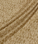 Woodcliff Dune fabric arranged in soft, flowing folds, showing its repeating botanical pattern of oak-inspired leaves and intertwining branches in warm sandy beige and taupe against a cream background, with a subtle woven texture.