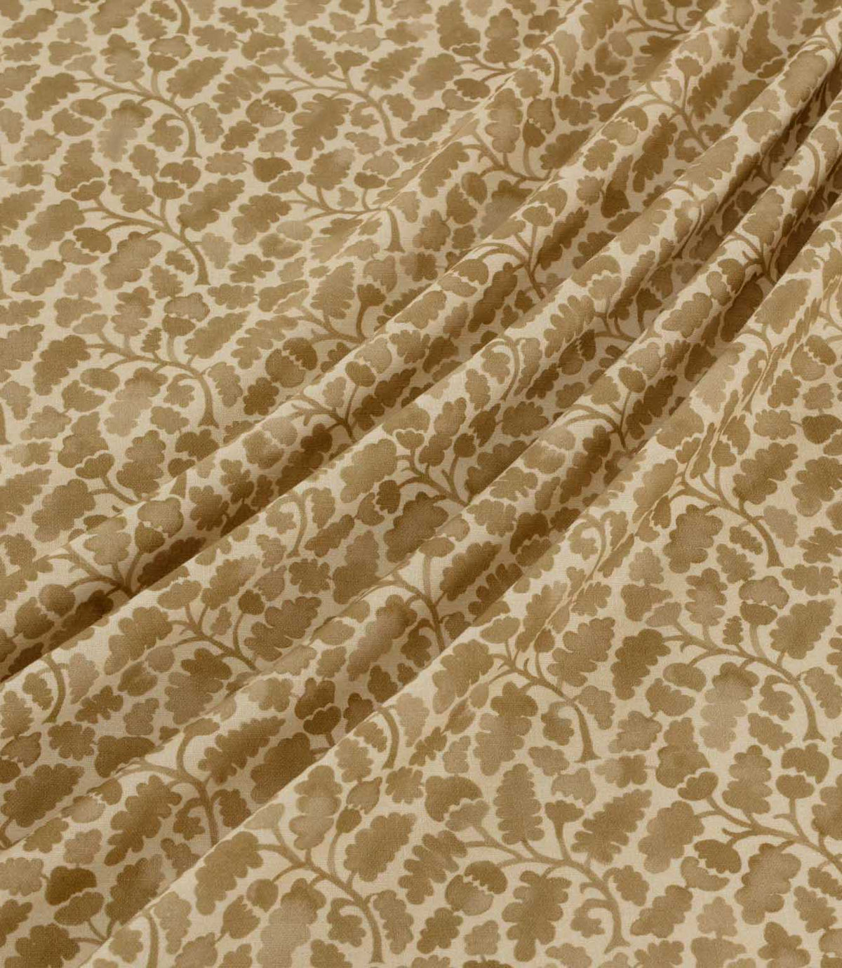 Woodcliff Dune fabric arranged in soft, flowing folds, showing its repeating botanical pattern of oak-inspired leaves and intertwining branches in warm sandy beige and taupe against a cream background, with a subtle woven texture.