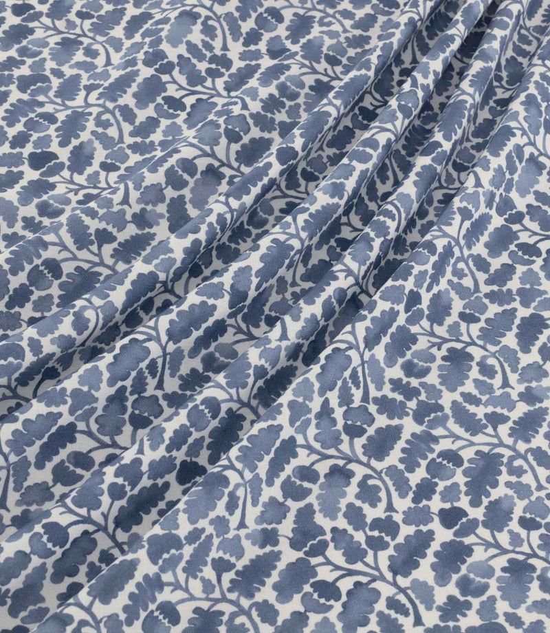 Woodcliff Nautica fabric arranged in soft, flowing folds, showcasing its repeating oak leaf and acorn design in navy and dusty blue tones on an ivory background, with a finely woven texture and delicate foliage detailing.