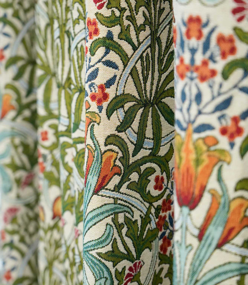 Close-up of Woodland Weeds Multi fabric featuring an intricate botanical design with green foliage, blue leaves and orange, red and yellow flowers on a natural cream background.