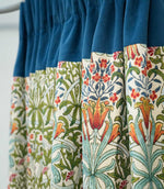 Woodland Weeds Multi fabric made into gathered curtains, featuring a colourful botanical pattern of green foliage, blue leaves and orange and red flowers, finished with a contrasting deep blue velvet heading.