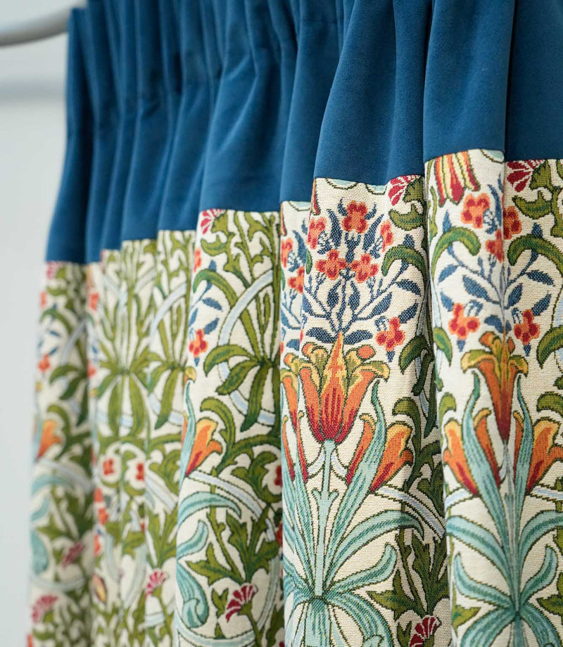 Woodland Weeds Multi fabric made into gathered curtains, featuring a colourful botanical pattern of green foliage, blue leaves and orange and red flowers, finished with a contrasting deep blue velvet heading.