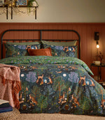 Woodland Ritual reversible duvet cover set in navy, featuring green, purple and rust trees with illustrated deer, foxes and owls, styled on a black metal bed with rust and green accessories.