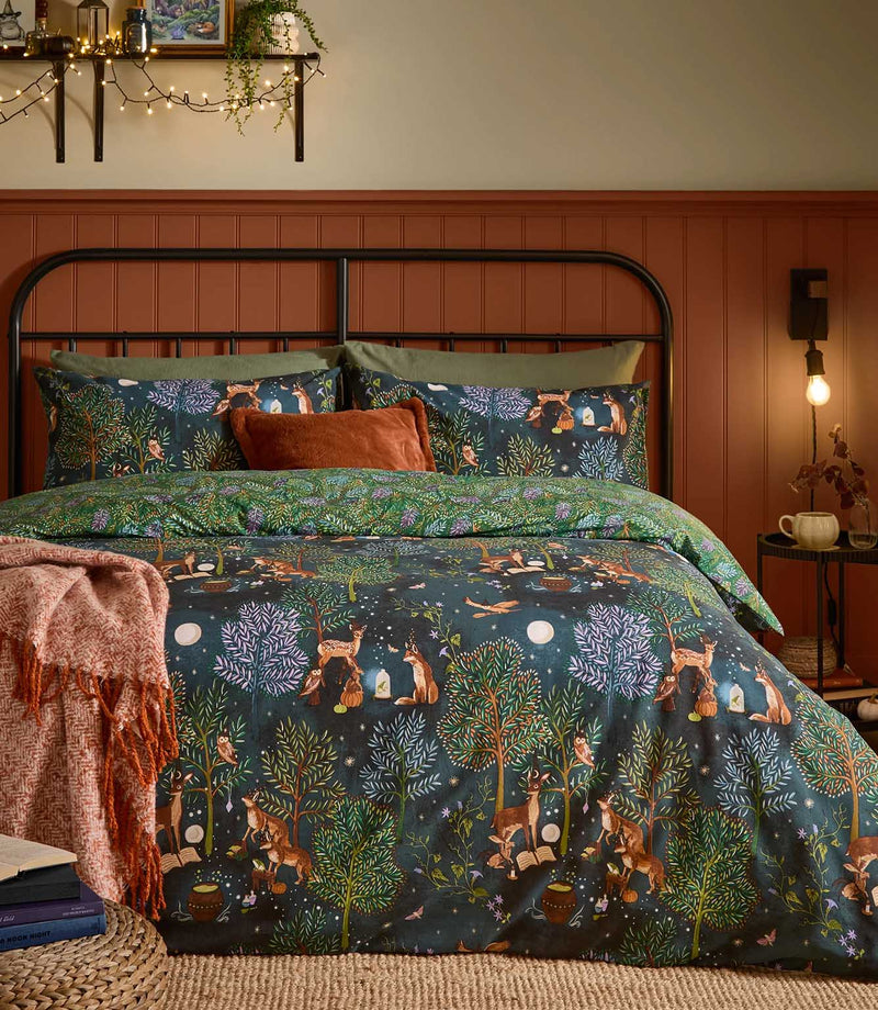 Woodland Ritual reversible duvet cover set in navy, featuring green, purple and rust trees with illustrated deer, foxes and owls, styled on a black metal bed with rust and green accessories.