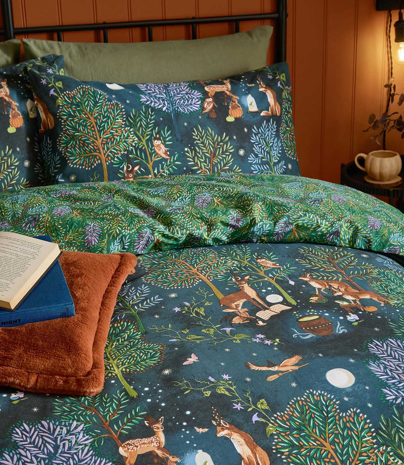 Close-up of the navy Woodland Ritual duvet cover and pillowcase, showing colourful woodland trees, deer, foxes, owls and celestial details with the green leafy reverse folded back.