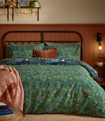 Woodland Ritual reversible duvet cover displayed on its green botanical reverse with dense blue, purple and rust foliage, with the navy woodland animal print folded back across the bed.