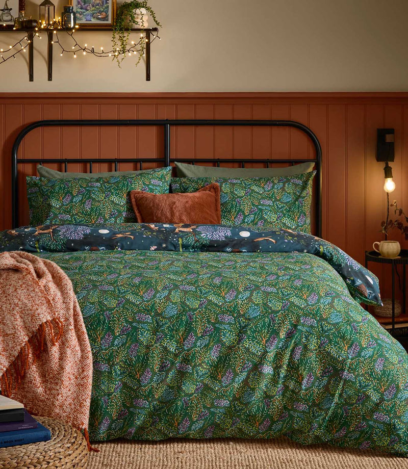 Woodland Ritual reversible duvet cover displayed on its green botanical reverse with dense blue, purple and rust foliage, with the navy woodland animal print folded back across the bed.